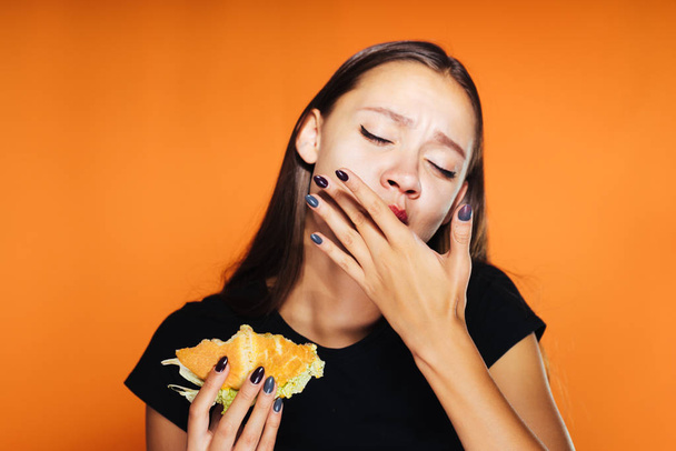 the hungry girl eats her hamburger. The girl licks her fingers. Very tasty food, isolated on an orange background - Photo, image