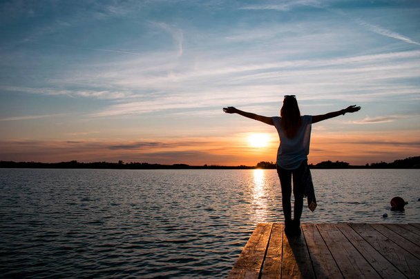 young woman standing with open arms on wooden pontoon in the lake and watching a colorful sunset - Foto, Bild