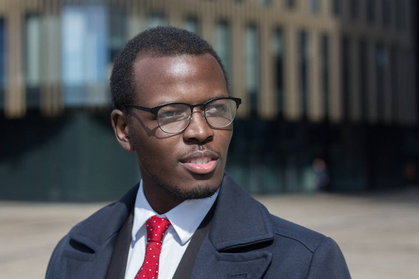 Retrato urbano de empresário afro-americano caminhando sozinho no centro da cidade vestido de casaco escuro e terno com gravata vermelha, olhando para rua com atenção e sorriso esperançoso em busca de novas ideias para o trabalho
 - Foto, Imagem