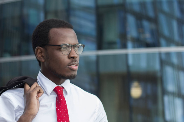 Horizontal image of dark-skinned African guy standing outdoors in white shirt with coat hanging on fingers, looking serious and tense as he is watching street in business city center during break - Photo, image