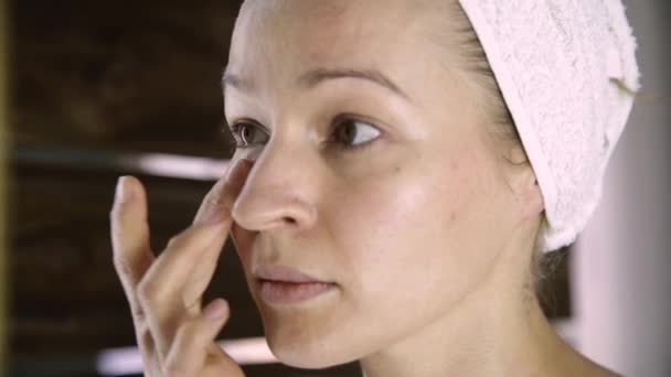 Beautiful healthy woman in a towel putting moisturizing cream on face. Skin care and home Spa - Materiaali, video