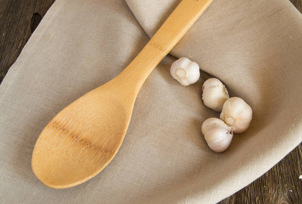 Garlic and a wooden spoon lying on a linen napkin. Old dark farm table, copy space - Photo, Image