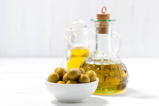 fles met olijfolie en kom met olijven op witte houten tafel - Foto, afbeelding