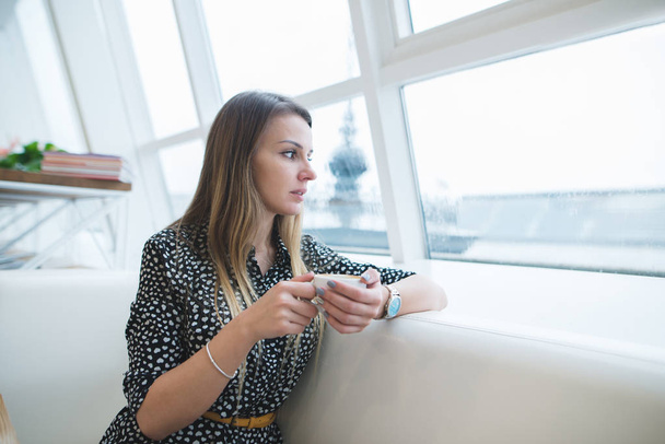 Een mooie vrouw met een kopje koffie in haar handen zit in een prachtig restaurant en kijkt uit het raam. De vrouw is warming-up met koffie in het café. - Foto, afbeelding