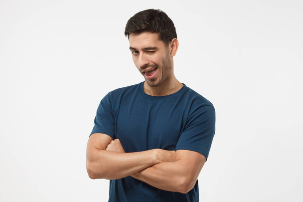 Handsome male with crossed arms smiling and winking, looking at camera isolated on gray background - Zdjęcie, obraz