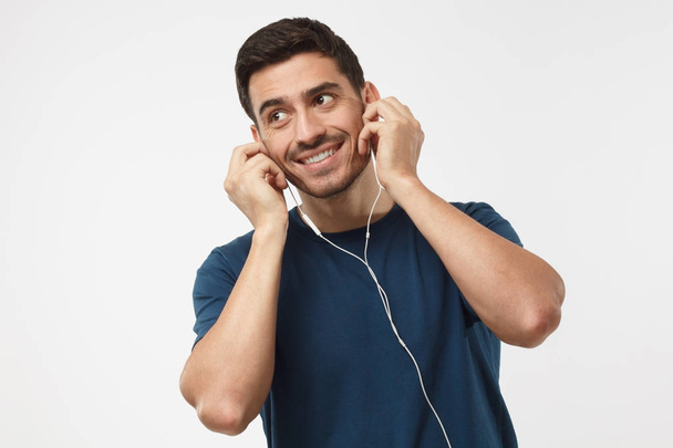 Portrait of happy young man in blue t-shirt, holding earphones with both hands, listening music, smiling, isolated on gray background - Fotografie, Obrázek