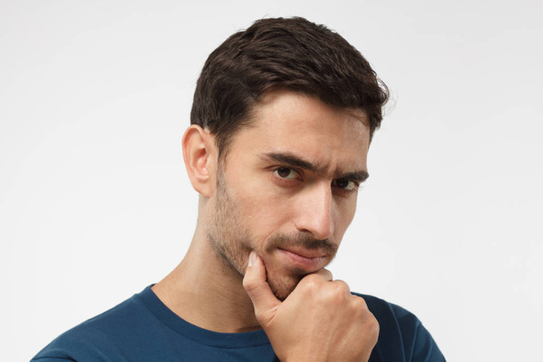 Close up portrait of young handsome male in blue t-shirt touching his chin with hand thoughtfully looking at camera. Doubt concept  - Foto, Imagem