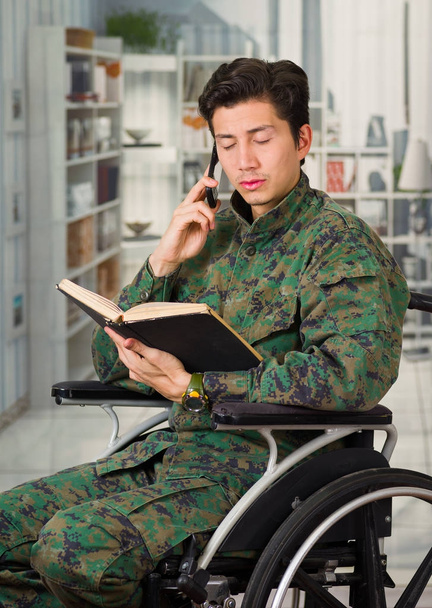 Nahaufnahme eines hübschen jungen Soldaten, der im Rollstuhl sitzt, zu Hause ein Buch liest und sein Handy vor verschwommenem Hintergrund benutzt - Foto, Bild