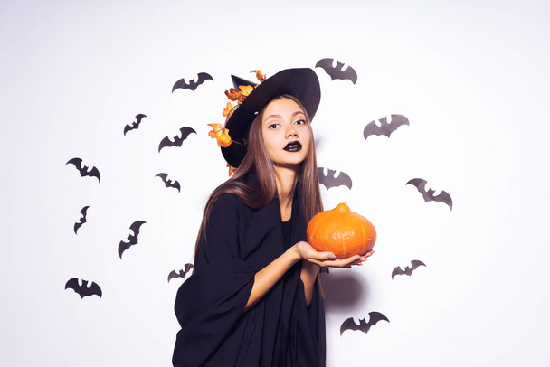a young girl with a black lipstick pretends to be a witch for Halloween, hold an orange pumpkin in her hand and looks gloomy - Photo, Image