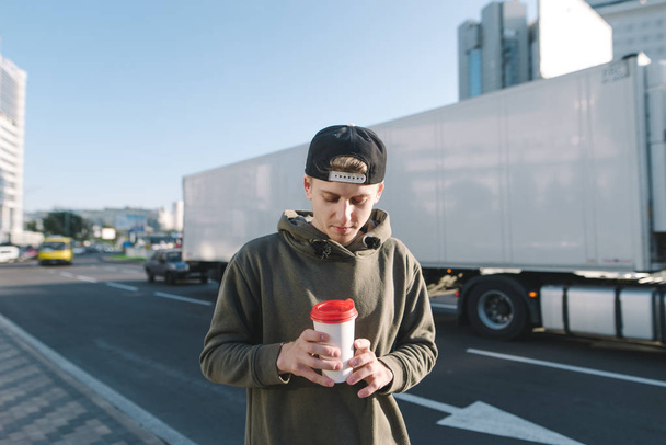 Ένας νεαρός, κομψό σπουδαστής στέκεται στο δρόμο και βλέπει ένα φλιτζάνι του καφέ στα χέρια του. Μαθητής στέκεται ενάντια σε μια άκρη του δρόμου και άσπρο φορτηγό. - Φωτογραφία, εικόνα