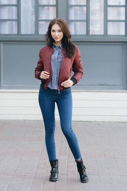 Young beautiful stylish woman in a burgundy pilots jacket and blue jeans, street style, spring and autumn trend, look, hair in the wind - Photo, image