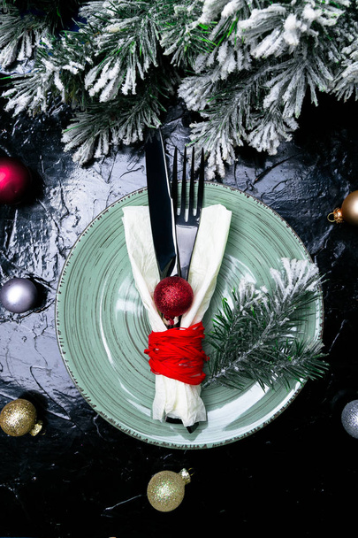 Vánoční prostírání stolu. Svátky na pozadí. Zelená deska, nůž a vidlička s vánoční výzdobou na černém stole. Pohled shora. - Fotografie, Obrázek