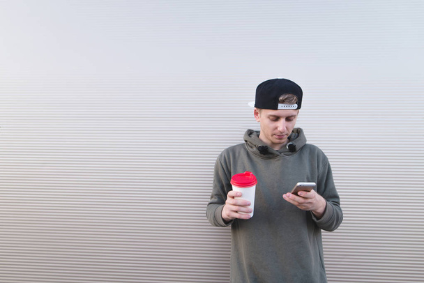 Κομψό νεαρό άνδρα με ένα φλιτζάνι καφέ στα χέρια του και βλέπουν ένα smartphone με φόντο έναν τοίχο φωτός. - Φωτογραφία, εικόνα