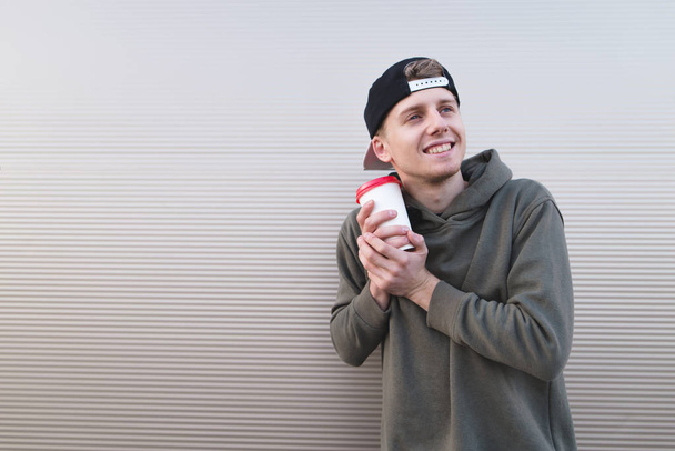 Ένα γλυκό και θετική νεαρός άνδρας με καφέ στα χέρια του αγκαλιάζει ένα φλιτζάνι με φόντο έναν τοίχο φωτός. - Φωτογραφία, εικόνα