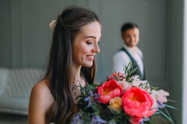 Close-up portrait of cheerful, young bride with a wedding bouquet on blurred groom background. A happy girl with a bunch of flowers - Zdjęcie, obraz
