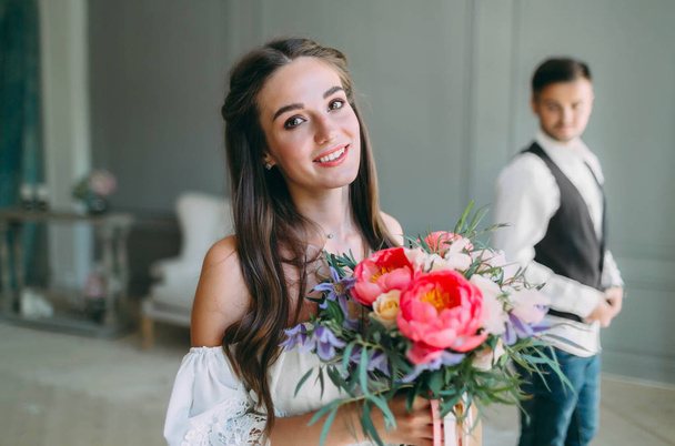 Close-up portrait of cheerful, young bride with a wedding bouquet on blurred groom background. A happy girl posing with a bunch of flowers - Photo, image