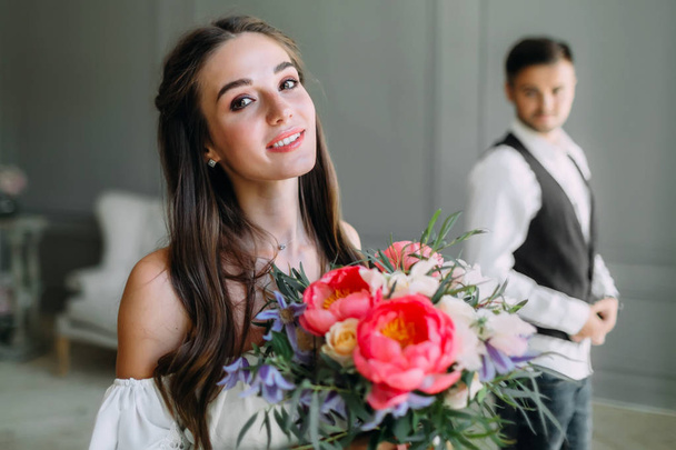 Close-up portrait of cheerful, young bride with a wedding bouquet on blurred groom background. A happy girl posing with a bunch of flowers - Photo, image
