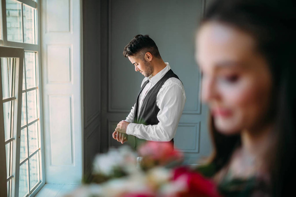 Handsome groom is preparing for wedding day on a background of blurred bride with a bouquet. Man in the festive clothes near a panoramic window. Artwork - Fotografie, Obrázek