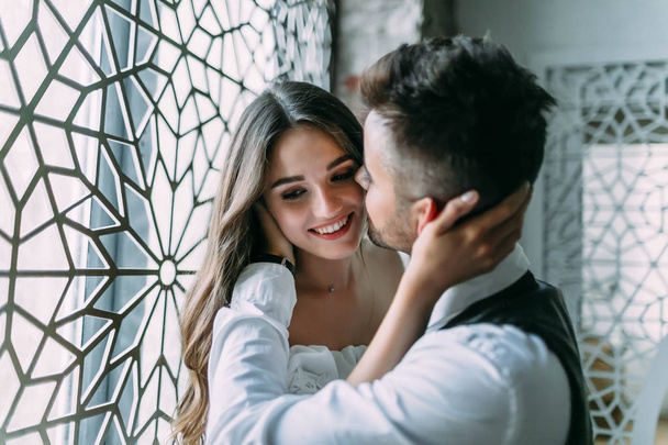 Cheerful young bride embraces grooms head while he kisses her in cheek on the vintage window background - Foto, afbeelding