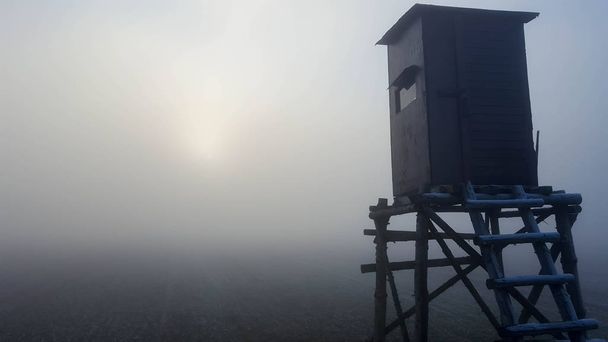 Paisagem melancólica de inverno nebuloso com um posto de vigia e campo durante o pôr do sol
 - Foto, Imagem
