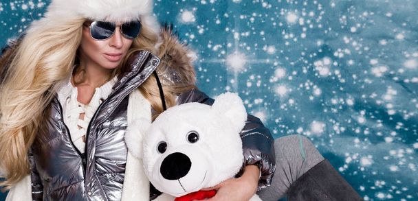 Beautiful stunning woman with long blond hair and perfect face dressed in winter clothing, silver warm jacket and fur cap and silver sunglasses on winter background - Photo, Image