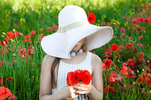 Suloinen tyttö hatussa, seisoo niityllä ja pitelee punaisia unikkoja.
 - Valokuva, kuva