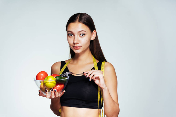 een atletische meisje horloges van haar figuur, bezit is een plaat met nuttig groenten en vruchten in haar handen - Foto, afbeelding