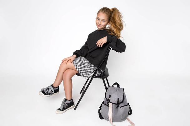 Cute little girl dressed in school clothes and school backpack sitting on white background - Photo, Image