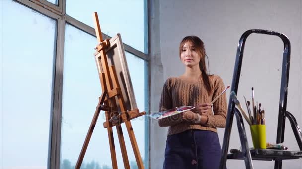 Giovane artista donna è in piedi vicino a un cavalletto e stand con strumenti di pittura
. - Filmati, video