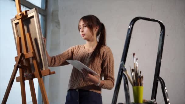 Young brunette is drawing by paintbrush, holding palette and standing near easel - Footage, Video