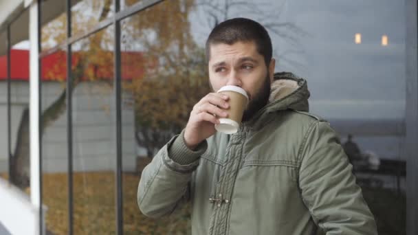 Όμορφος νεαρός γενειοφόρος άνδρας κρατά ένα κύπελλο. πίνοντας ζεστό ρόφημα καφέ ή τσάι σε εξωτερικούς χώρους το φθινόπωρο - Πλάνα, βίντεο