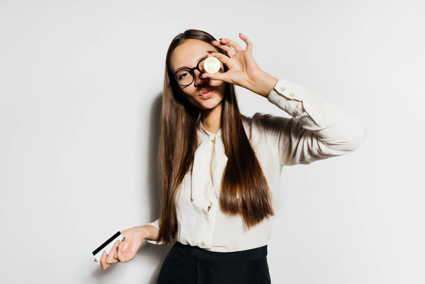 a nice girl with glasses is holding a bank card in her hand and attaching a gold coin to her eye. Bitcoins, crypto currency, electronic money. - Foto, imagen