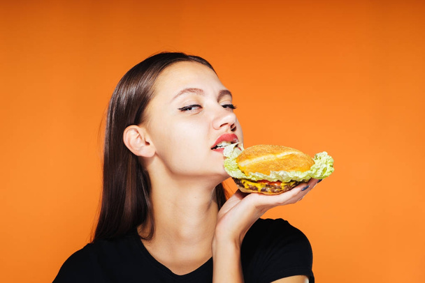 Sexy girl starts to eat her burger and looks directly into the camera. Isolated on orange background, hungry girl. - Photo, image