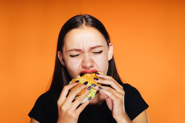 The girl greedily eats a burger. tasty food, isolated on orange background - Foto, afbeelding