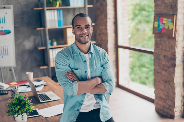 Portrait of successful black guy, looking at the camera, standing with crossed arms at his work place in the office, in casual smart, smiling - Fotó, kép