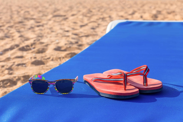 Pantoffels en zonnebril op zonnebank op strand - Foto, afbeelding