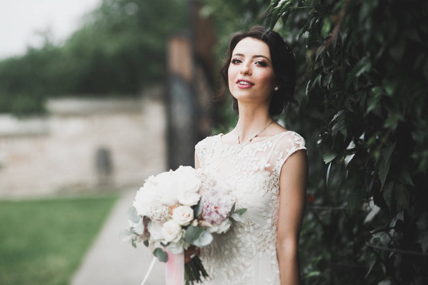 Beautiful brunette bride in elegant white dress holding bouquet posing neat trees - Фото, изображение