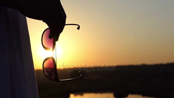 een jonge slanke vrouw staat met zonnebril in haar handen bij zonsondergang in Slo-Mo - Video