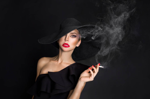 Elegáns nő, femme fatale, fekete kalap cigarettával a kezében. Fekete háttér - Fotó, kép