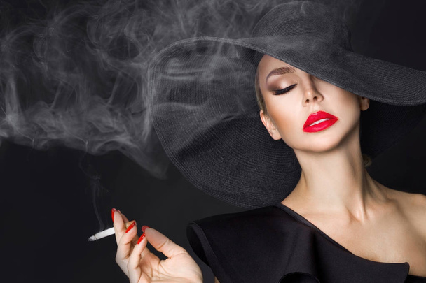 Elegant woman, femme fatale in black hat with cigarette in hand. On black background - Foto, afbeelding