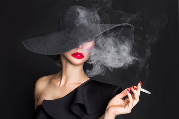 Elegant woman, femme fatale in black hat with cigarette in hand. On black background - Valokuva, kuva