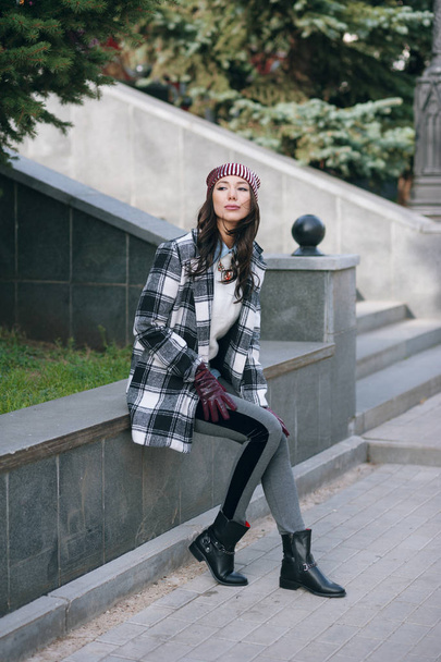 Young beautiful stylish woman, wearing a coat in a white and black cage, white jacket, gray trousers, black boots. Street style, spring and autumn trend, a flirty look - Valokuva, kuva