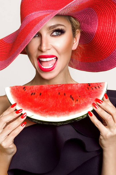 Beautiful female model with perfect face and red hat holding a watermelon at her face - Photo, Image
