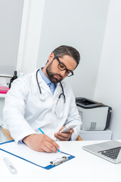 doctor using smartphone and writing in clipboard - Photo, Image