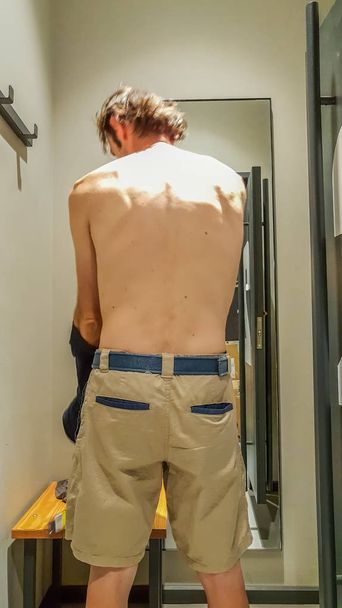 Rear view of a young Caucasian handsome man trying new clothes in front of a mirror in a changing room of a clothing store - Photo, Image