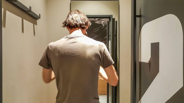 Visão traseira de um jovem caucasiano bonito homem tentando roupas novas na frente de um espelho em um vestiário de uma loja de roupas
 - Foto, Imagem