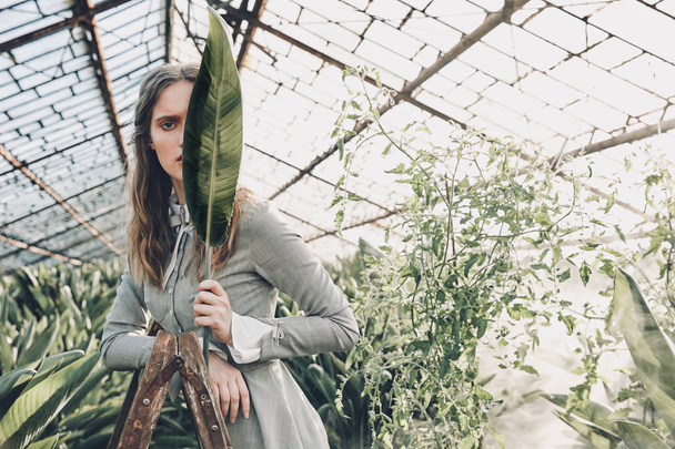 Young beautiful girl with long brown hair wearing light gray dress in retro style, posing with long green leaf in greenhouse, full of plants - Foto, afbeelding
