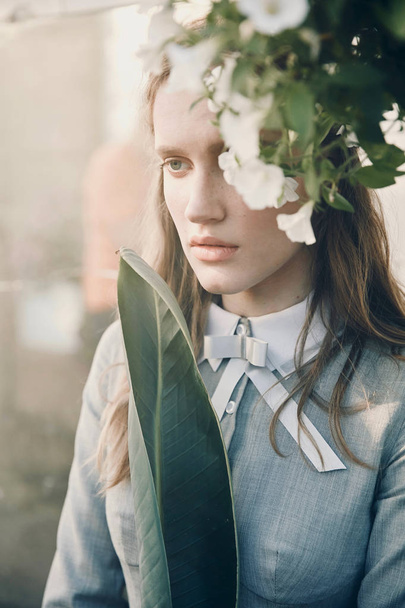 Portrait of young beautiful serious girl with long brown hair wearing light gray dress in retro style, posing near white flowers in greenhouse full of plants - Photo, Image