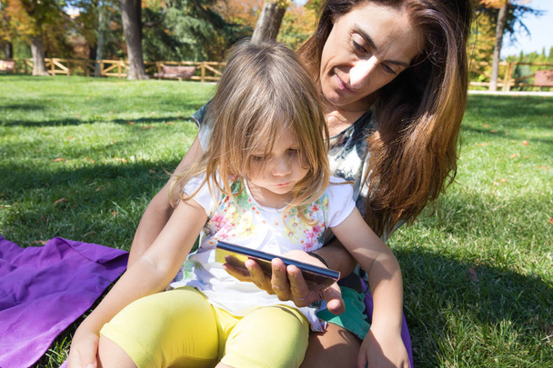 маленька дівчинка і мати дивиться мобільний, сидячи на траві
 - Фото, зображення