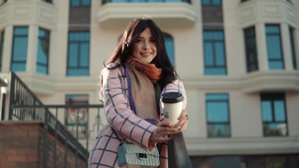 Portrait of a cute young woman against the backdrop of an autumn city. a girl in an autumn coat drinks coffee from a paper cup and enjoys a warm autumn day - Πλάνα, βίντεο
